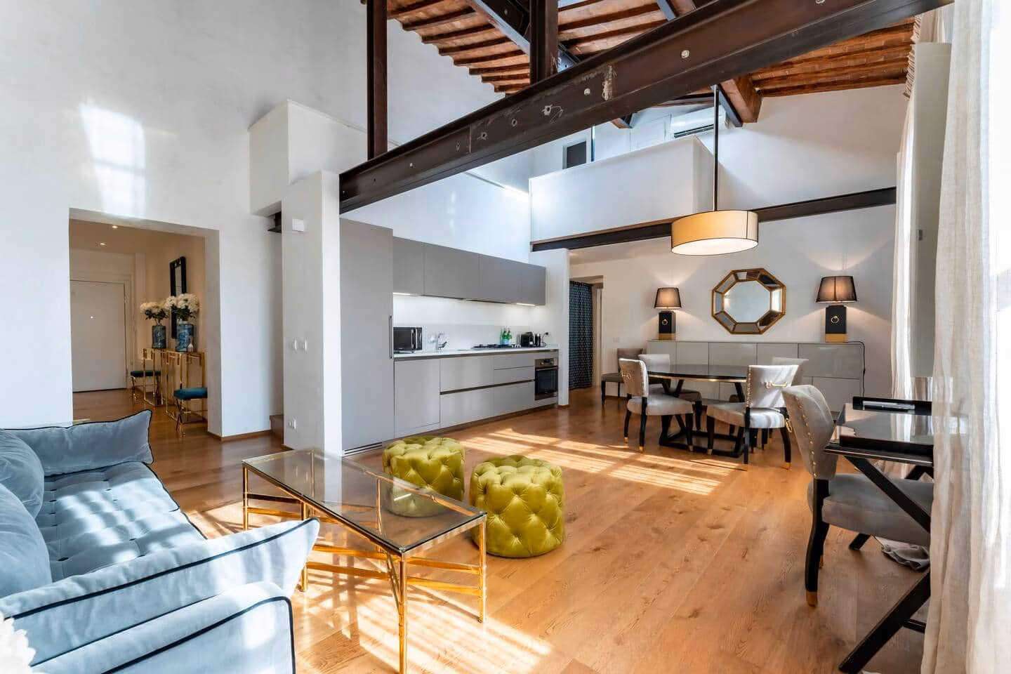 At Home — light-filled open-plan living and dining space beneath a double-height beamed ceiling, with a pale-blue sofa